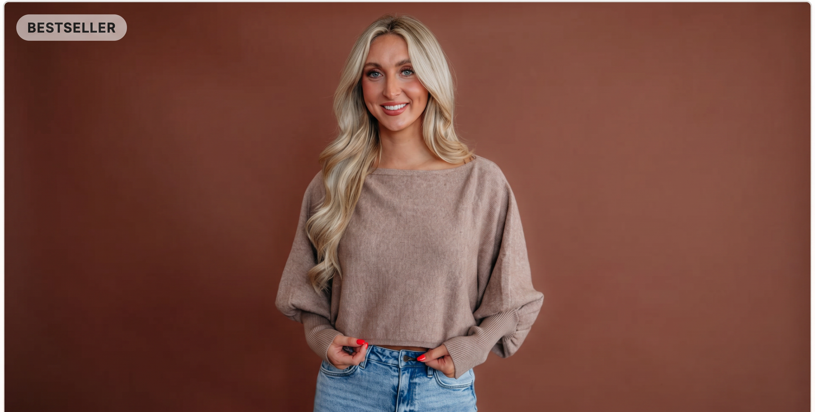 A woman with long blonde hair wearing a beige wide-neck knit sweater and blue jeans, standing against a solid brown studio background.